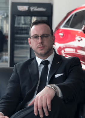 Man in a suit sitting confidently with a red car in the background.