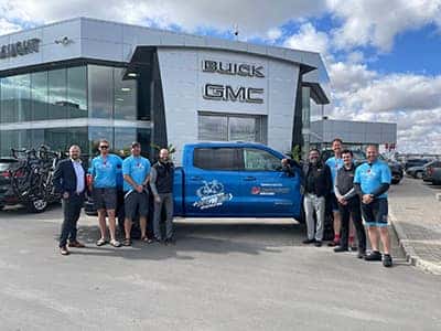 Group of people stand with a blue truck outside a Buick GMC dealership, under a partly cloudy sky.