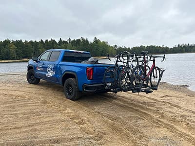 A blue pickup truck with a bike rack carrying three bicycles parked by a lakeside beach under a cloudy sky.