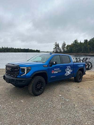 Blue truck with logos parked by a lake, forested backdrop, bicycles in the bed.