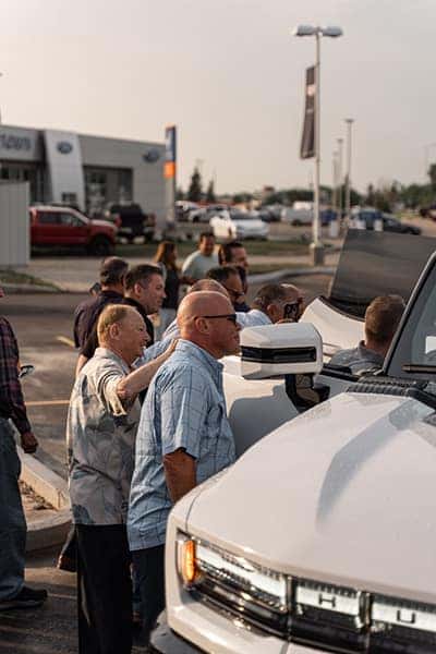 A group of people examines white trucks at a dealership under a clear sky.