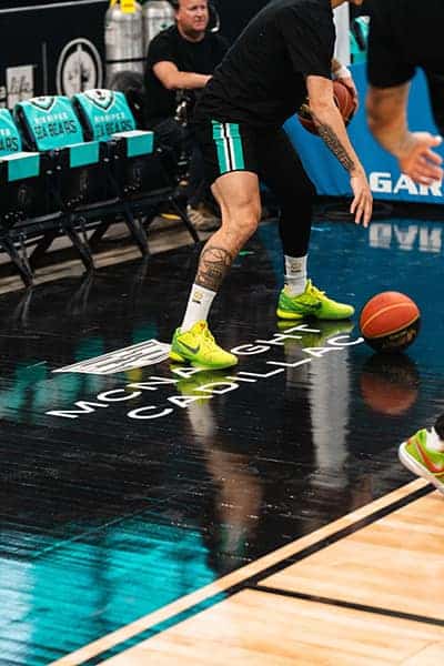 Basketball player dribbling on a court with "MCW MIGHT CADILLAC" visible, wearing bright green shoes.