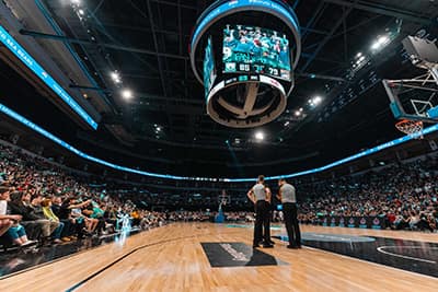 Basketball game in a packed arena, referees discussing under scoreboard, vibrant atmosphere.