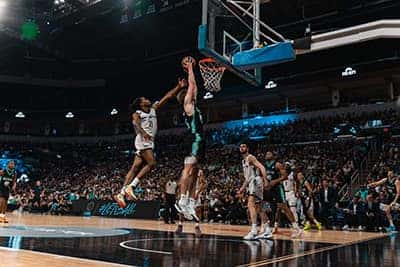 Basketball player attempts a layup during a game, with a defender closely contesting.