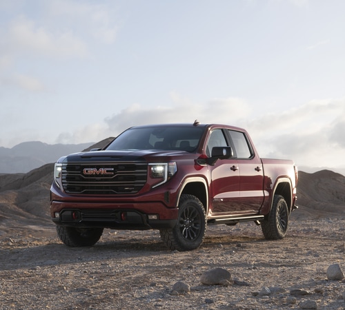 Red GMC pickup truck parked on a rocky terrain with mountainous backdrop under a cloudy sky.