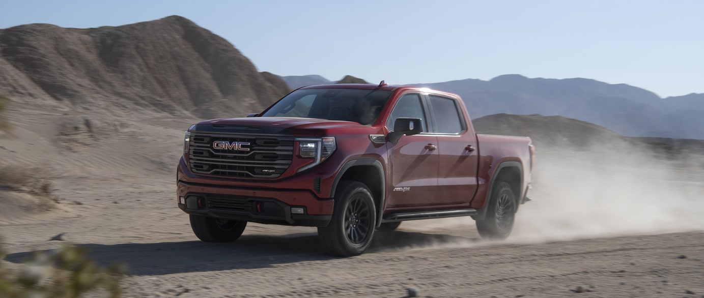 A green SUV is parked on a paved surface near a calm body of water. The car features a prominent grille and sleek design. In the background, hills are visible under a clear blue sky.