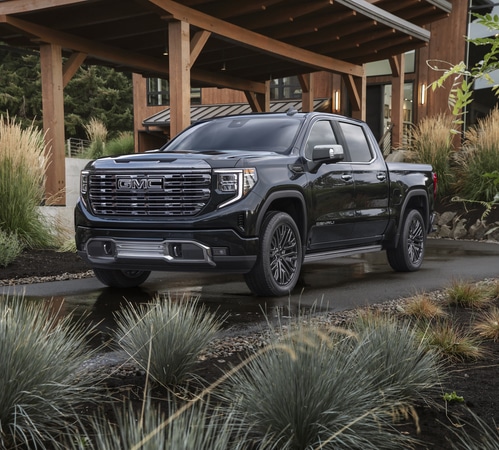 Black GMC Sierra truck parked in front of a modern wooden house with lush greenery.