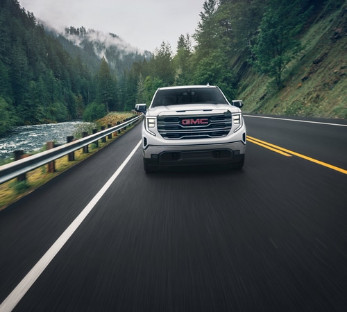 GMC truck driving on a scenic forested road alongside a river, showcasing rugged landscape.