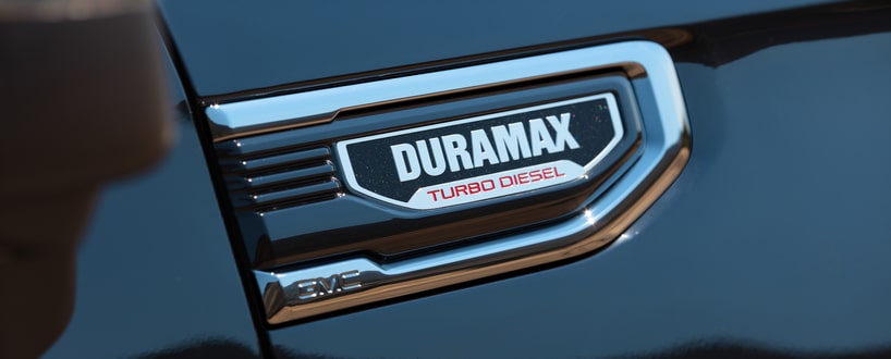 A person in a pink top and blue pants stands behind a green SUV parked on the side of a road. Theyre reaching towards the cars back. A grassy park with trees and people is visible in the background under a clear sky.