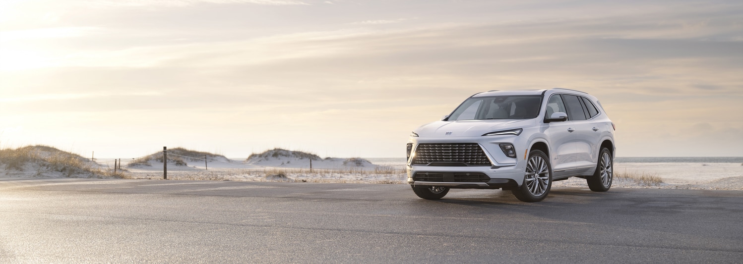 A shiny red SUV is parked in a dimly lit urban setting at night. The car features a sleek design with distinctive wheel rims, and illuminated headlights, reflecting light on the wet pavement. Modern architectural elements are visible in the background.