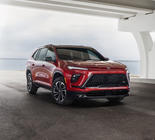 Red SUV parked under a modern structure near the ocean, showcasing sleek design and grille.