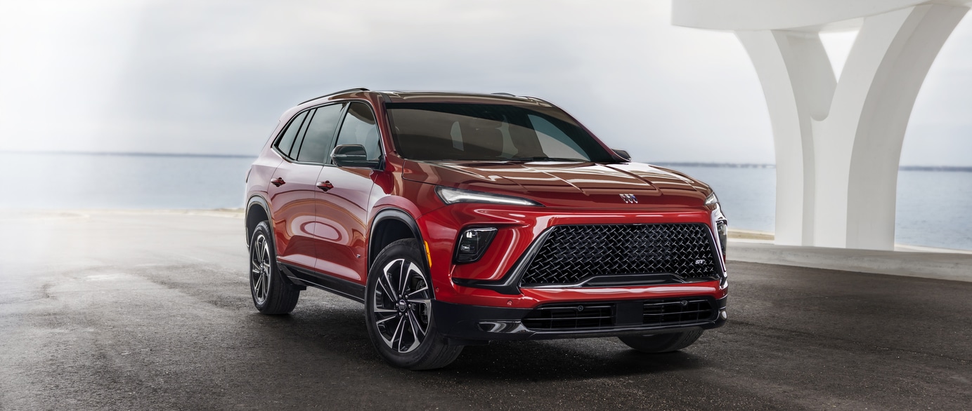 A green SUV is parked on a paved surface near a calm body of water. The car features a prominent grille and sleek design. In the background, hills are visible under a clear blue sky.