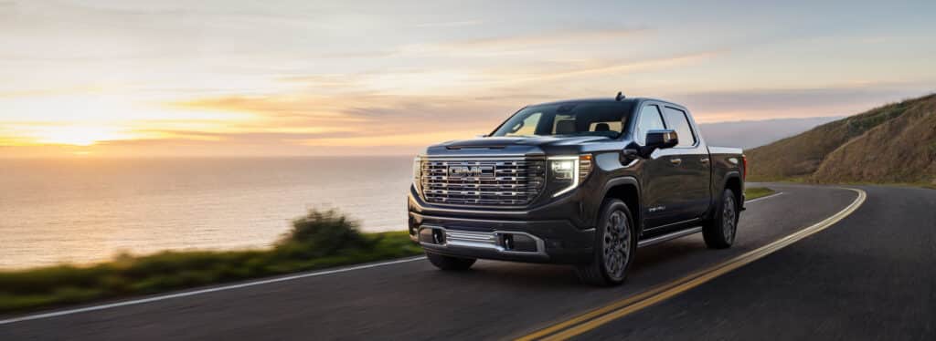 Black GMC truck driving on a coastal road at sunset, with ocean views on the left.