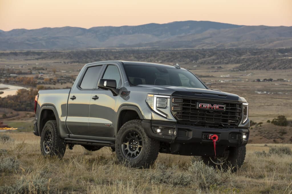 Silver GMC truck parked in a grassy field with mountain range backdrop at sunset.