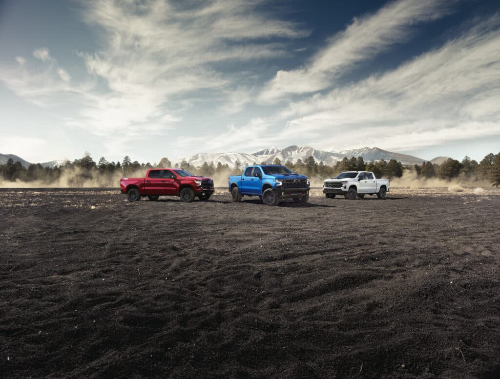 Red, blue, and white trucks on a rugged landscape with mountains and trees in the background.