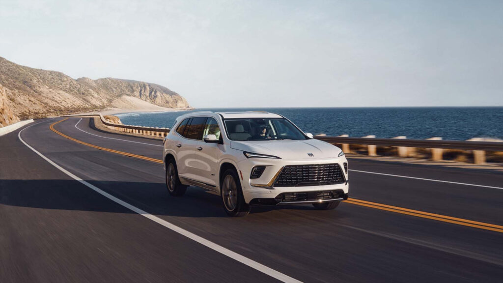 White SUV driving along a scenic coastal highway with ocean and cliffs in the background.