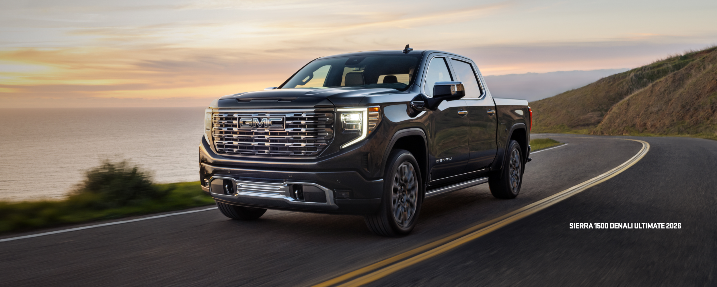 GMC Sierra 1500 Denali Ultimate 2026 driving on a coastal road at sunset.