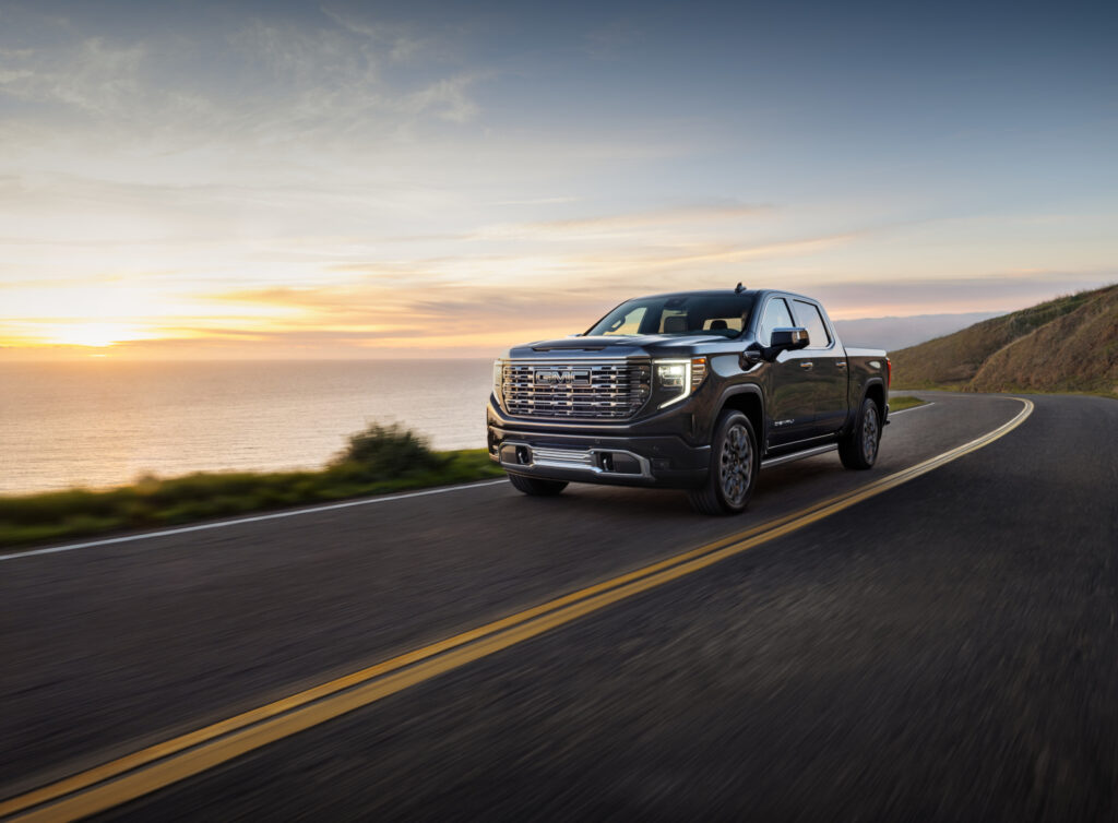 Pickup truck driving on a coastal road at sunset, with ocean and hills in the background.