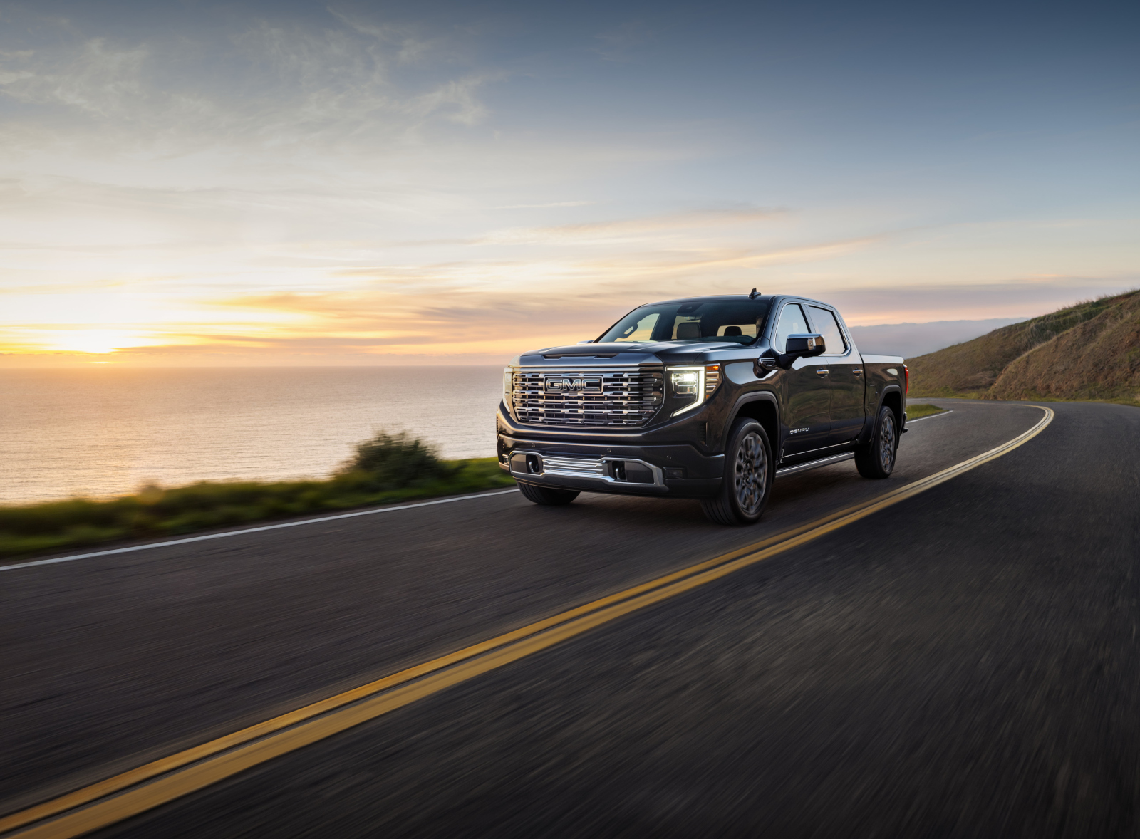 Pickup truck driving on a coastal road at sunset, with ocean and hills in the background.